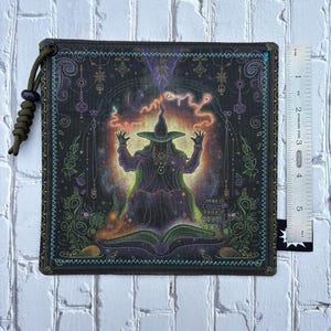 May include: Square, dark green fabric patch with a detailed illustration of a witch in a pointed hat, surrounded by mystical elements. The witch is seated in front of an open book, with a ruler next to the patch. The patch has a green cord loop.
