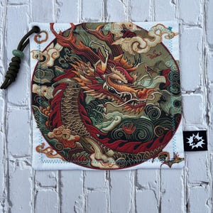 May include: Square white pouch featuring a detailed illustration of a red and gold dragon within a circular design. The dragon is surrounded by clouds and intricate patterns. A black tag with a white graphic is on the bottom right.