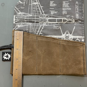 K&ouml;nnte beinhalten: Eine braune Stiftetui aus Canvas mit vier F&auml;chern und einem Lineal mit einer L&auml;nge von 17,8 cm. Die Etui ist mit einem Stoff verziert, der eine Schwarzwei&szlig;illustration eines McDonnell Douglas F/A-18A Hornet Kampfjets zeigt.