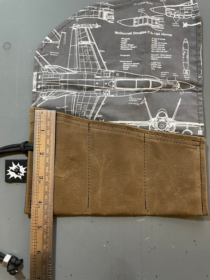 K&ouml;nnte beinhalten: Eine braune Stiftetui aus Canvas mit vier F&auml;chern und einem Lineal mit einer L&auml;nge von 17,8 cm. Die Etui ist mit einem Stoff verziert, der eine Schwarzwei&szlig;illustration eines McDonnell Douglas F/A-18A Hornet Kampfjets zeigt.
