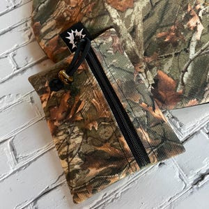 May include: A small camouflage-patterned pouch with a zipper closure and a drawstring. The pouch has a black zipper and a black drawstring. The pouch is made of a fabric with a camouflage pattern.