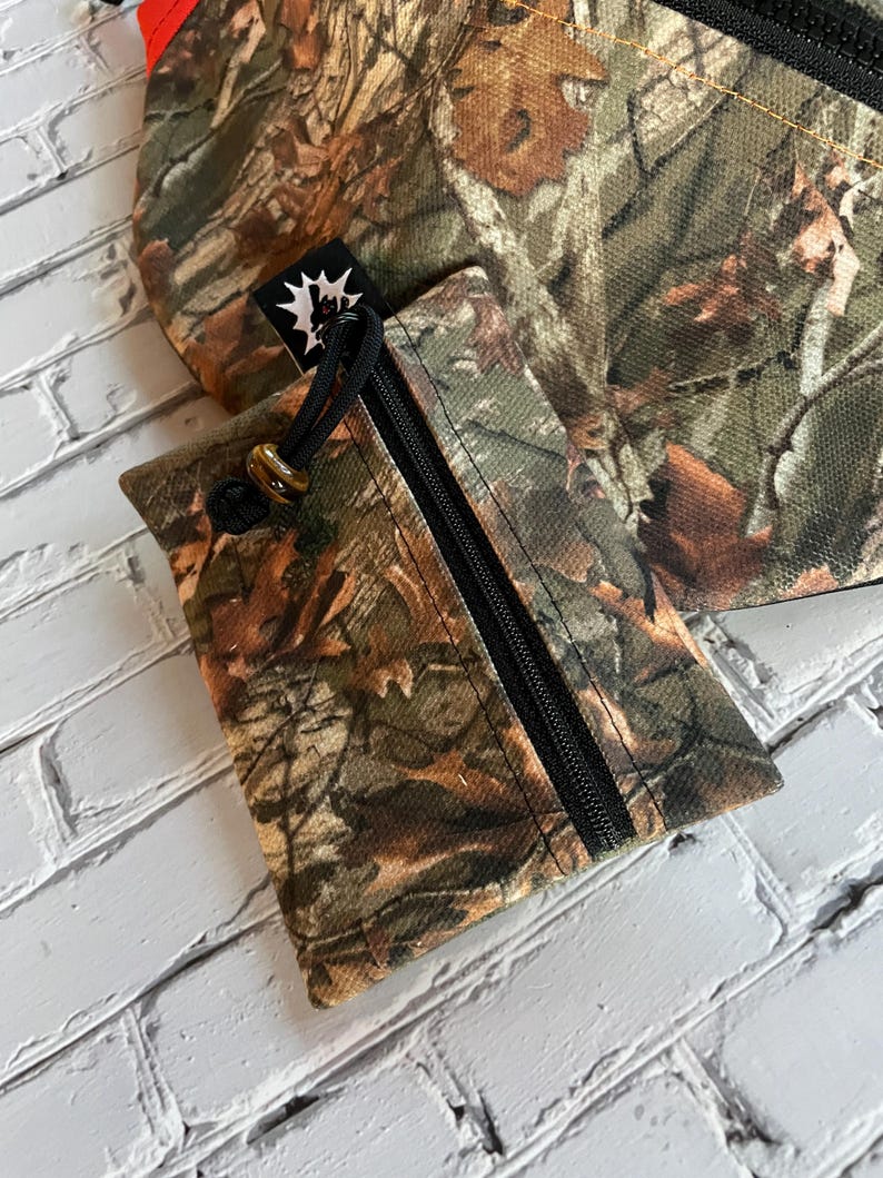 May include: A small camouflage-patterned pouch with a zipper closure and a drawstring. The pouch has a black zipper and a black drawstring. The pouch is made of a fabric with a camouflage pattern.