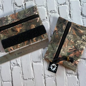 May include: Two camouflage zippered pouches with black zippers and black velcro closures. The pouches are made of a fabric with a camouflage pattern. One pouch has a ruler next to it.