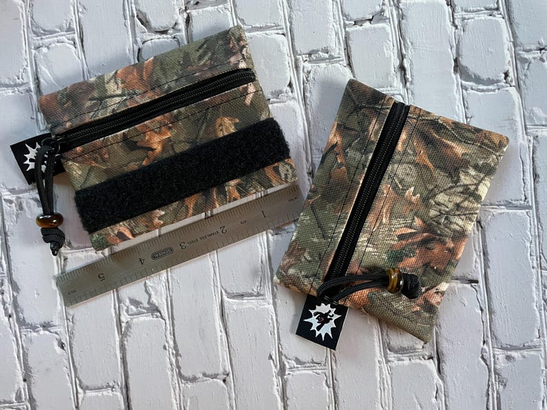 May include: Two camouflage zippered pouches with black zippers and black velcro closures. The pouches are made of a fabric with a camouflage pattern. One pouch has a ruler next to it.