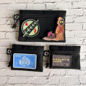May include: Three black pouches with zip closures and various patches. One pouch features a green and red emblem, another a Jawa character, and the third a blueprint design. The last pouch has the text "LV-426 HADLEY'S HOPE".