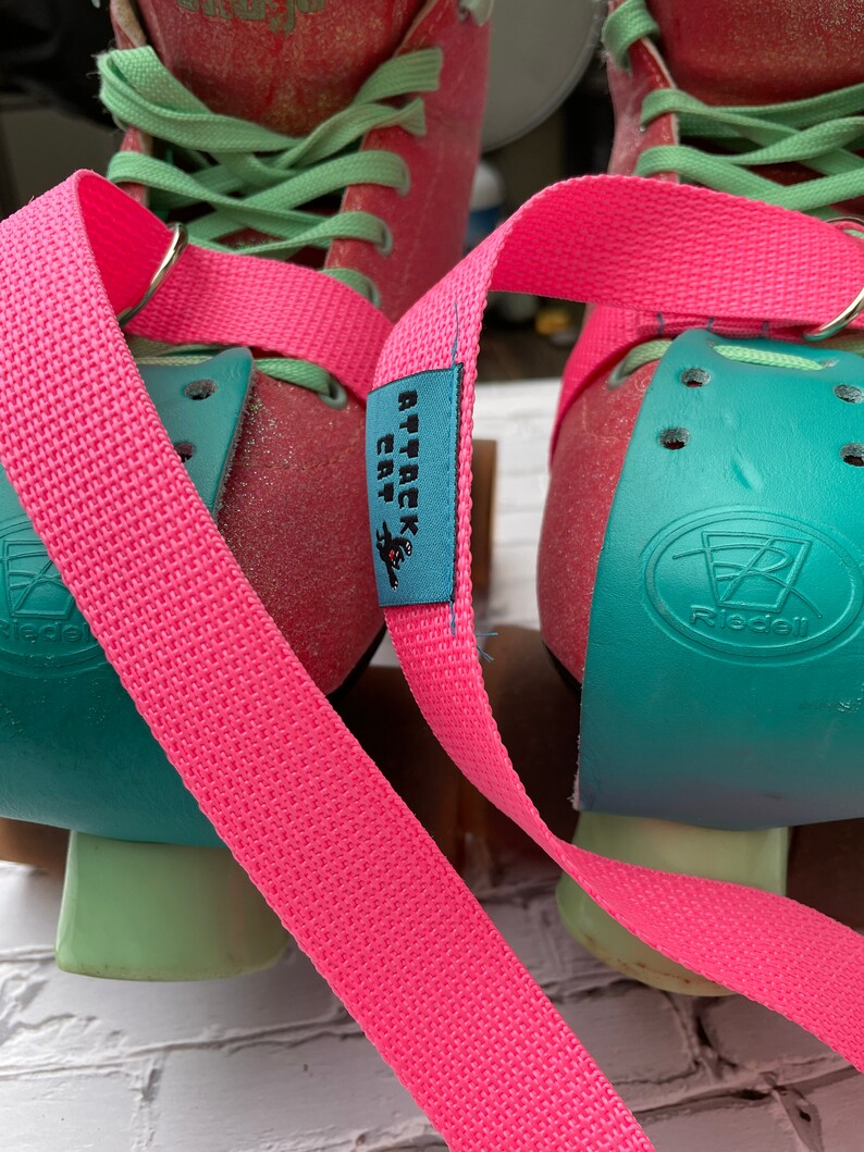 Puede incluir: Un par de patines de ruedas rosas con topes de dedos de color turquesa y cordones verdes. Los patines tienen correas rosas con una etiqueta azul que dice "Attack Cat".