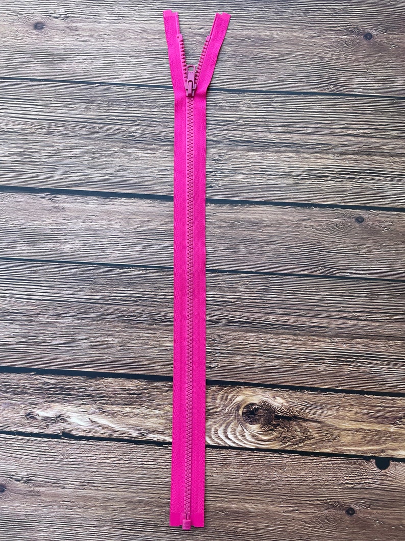 May include: A pink zipper with a metal slider. The zipper is lying flat on a wooden surface.