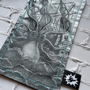May include: A black and white graphic print of an octopus attacking a ship. The print is on a white fabric pouch with a blue stitched border. The pouch has a black tag with a white graphic of a creature with a speech bubble.