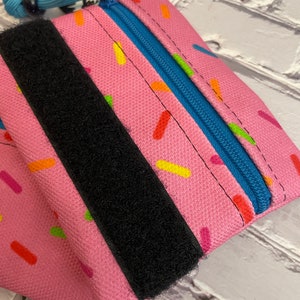 May include: A pink zippered pouch with a black velcro strap and a blue zipper. The pouch has a sprinkle pattern.