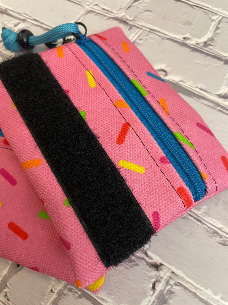 May include: A pink zippered pouch with a black velcro strap and a blue zipper. The pouch has a sprinkle pattern.