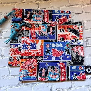 May include: A drawstring pouch featuring a vibrant Japanese-inspired print with red, blue, and black panels. The fabric displays illustrations of fish, crabs, and stylized waves, accented by Japanese kanji characters and a bright teal cord closure.
