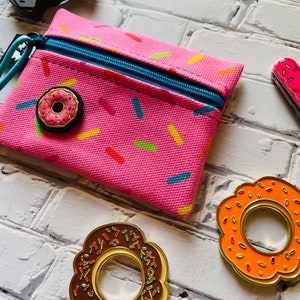 May include: A pink zippered pouch with a donut pattern and a donut-shaped zipper pull. There are three donut-shaped pins, one with chocolate frosting, one with orange frosting, and one with pink frosting.