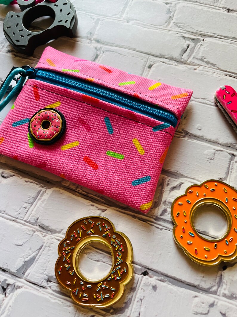 May include: A pink zippered pouch with a donut pattern and a donut-shaped zipper pull. There are three donut-shaped pins, one with chocolate frosting, one with orange frosting, and one with pink frosting.