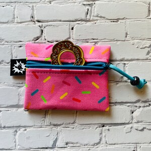 May include: A pink zippered pouch with a donut-shaped teething toy inside. The pouch has a sprinkle pattern and a blue zipper pull with a black bead.