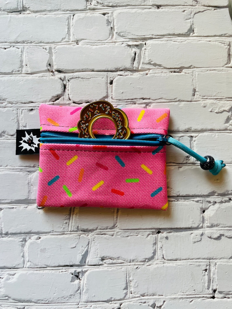 May include: A pink zippered pouch with a donut-shaped teething toy inside. The pouch has a sprinkle pattern and a blue zipper pull with a black bead.