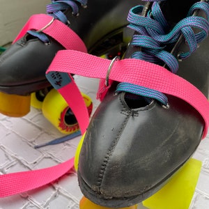 Puede incluir: Patines de ruedas de cuero negro con ruedas amarillas y correas rosas. Los cordones son de color turquesa y morado.