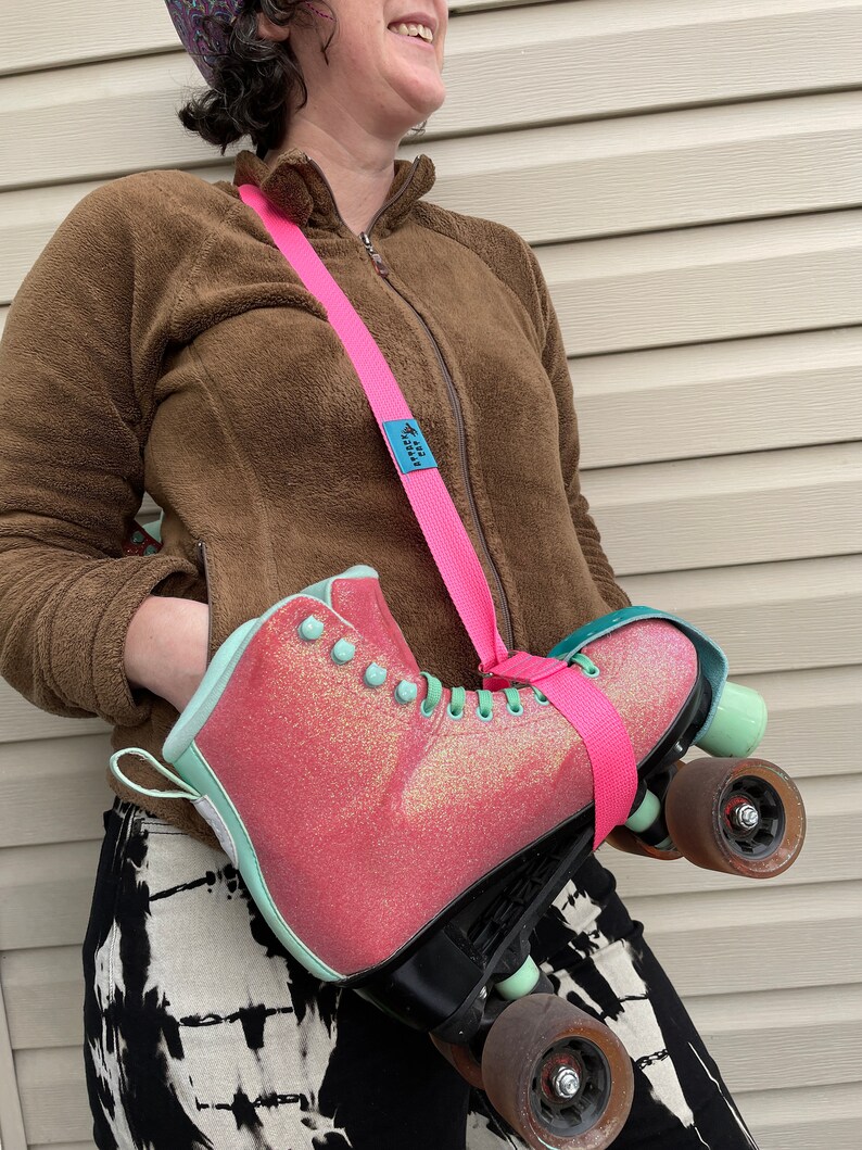 Puede incluir: Un pat&iacute;n de ruedas rosa y verde con purpurina en la bota est&aacute; siendo llevado por una persona usando una correa rosa y azul. La correa tiene el texto "Roll With It".