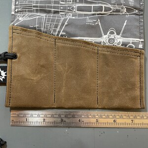 K&ouml;nnte beinhalten: Eine braune Werkzeugrolle aus Canvas mit drei Taschen. Die Werkzeugrolle ist aus strapazierf&auml;higem Material gefertigt und hat ein gen&auml;htes Design. Die Werkzeugrolle liegt auf einer grauen Oberfl&auml;che mit einem Lineal daneben.