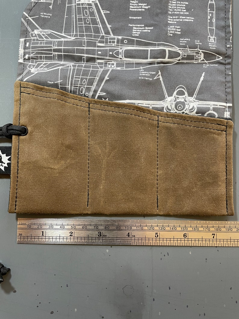 K&ouml;nnte beinhalten: Eine braune Werkzeugrolle aus Canvas mit drei Taschen. Die Werkzeugrolle ist aus strapazierf&auml;higem Material gefertigt und hat ein gen&auml;htes Design. Die Werkzeugrolle liegt auf einer grauen Oberfl&auml;che mit einem Lineal daneben.