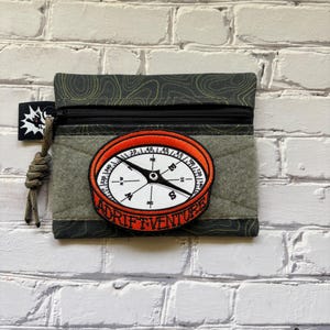 May include: A dark green zippered pouch featuring a topographic map pattern and a central circular patch depicting a compass with an orange border and the text ADRIFT VENTURE. A black zipper pull with a knotted olive green cord is attached to the side.