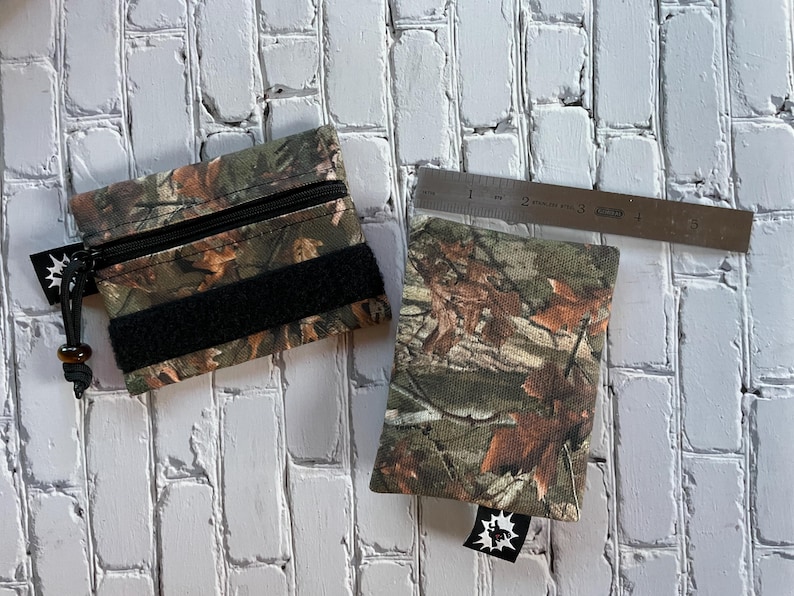 May include: Two small zippered pouches with a camouflage pattern. The pouches have black straps and a small black tag with a white starburst design.