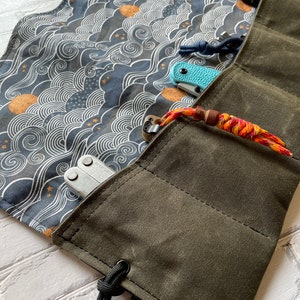 Pode incluir: Uma bolsa de lona verde que se enrola com vários bolsos para guardar ferramentas ou facas. A bolsa tem um fecho de cordão e uma alça preta com um logotipo branco. O interior da bolsa é forrado com um tecido azul com um padrão de ondas brancas.