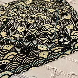Könnte beinhalten: Ein schwarzer und weißer Stoff mit einem sich wiederholenden Muster aus stilisierten Katzen und Wellen. Der Stoff hat einen blauen, genähten Rand.