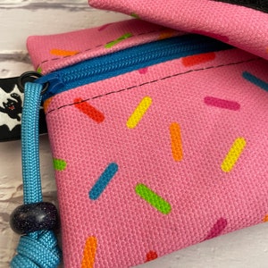 May include: Pink zippered pouch with a sprinkle pattern and a blue drawstring. The pouch has a black zipper pull and a blue bead on the drawstring.