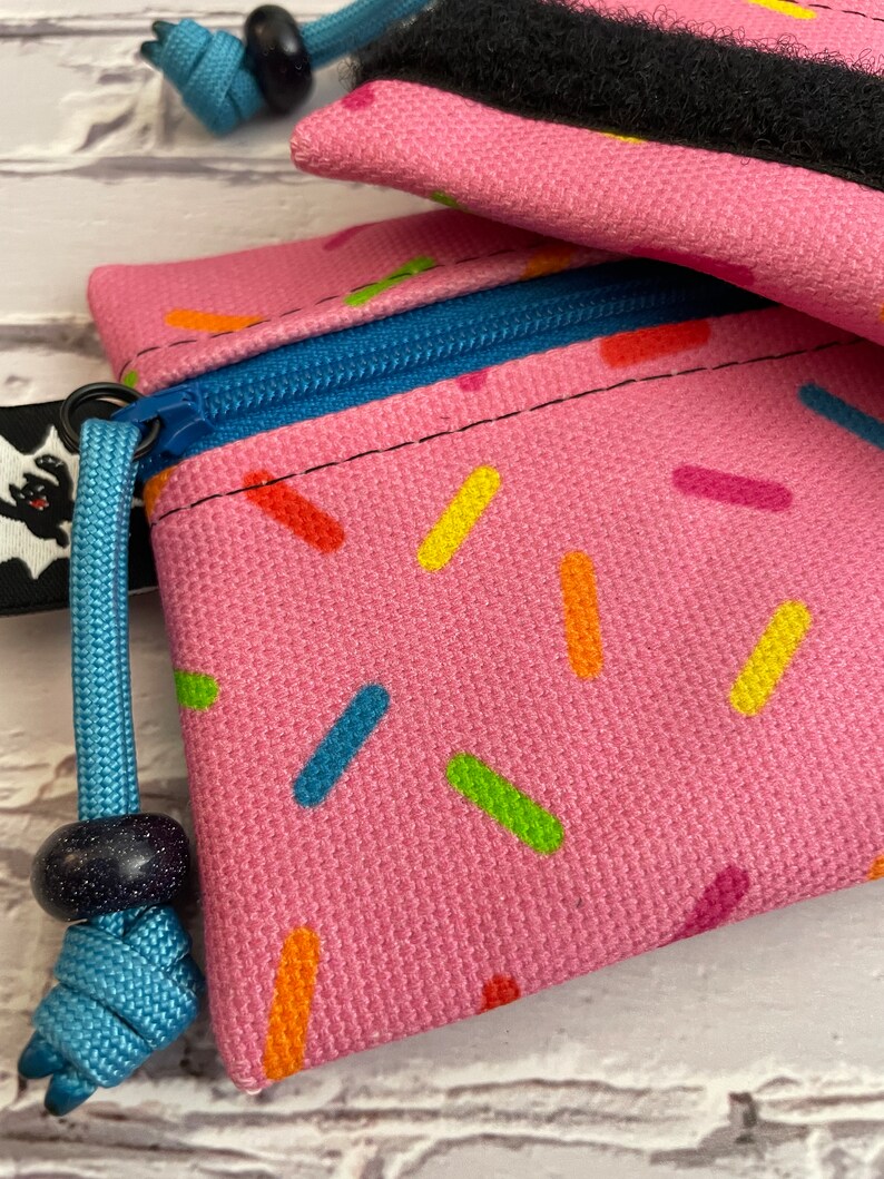 May include: Pink zippered pouch with a sprinkle pattern and a blue drawstring. The pouch has a black zipper pull and a blue bead on the drawstring.