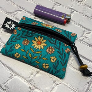 Puede incluir: Bolsa con cremallera floral turquesa con cremallera negra y cordón negro con una cuenta amarilla. Un encendedor Bic morado está al lado de la bolsa. La bolsa tiene una etiqueta negra con un gráfico blanco.