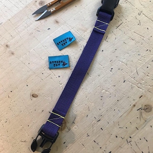 May include: A purple strap with a black plastic buckle and yellow stitching. Two blue fabric labels with the text "Attack Cat" are visible in the background.