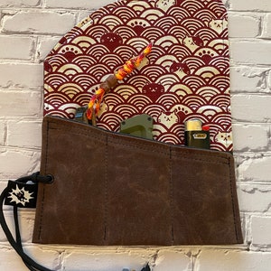 May include: A brown canvas roll-up pouch with a drawstring closure and a flap with a red and white cat pattern. The pouch has multiple slots for carrying small items like knives, lighters, and other tools.