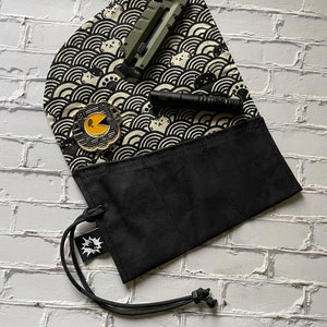 K&ouml;nnte beinhalten: Eine schwarze Stofftasche mit Kordelzug und einer kleinen Tasche auf der Vorderseite. Die Tasche hat drei F&auml;cher im Inneren. Die Tasche liegt auf einer wei&szlig;en Ziegelstein-Oberfl&auml;che. Die Tasche ist mit einem schwarz-wei&szlig;en Muster aus Wellen und Katzen verziert. Ein gr&uuml;nes Universalmesser und eine schwarze Taschenlampe liegen auf der Tasche.