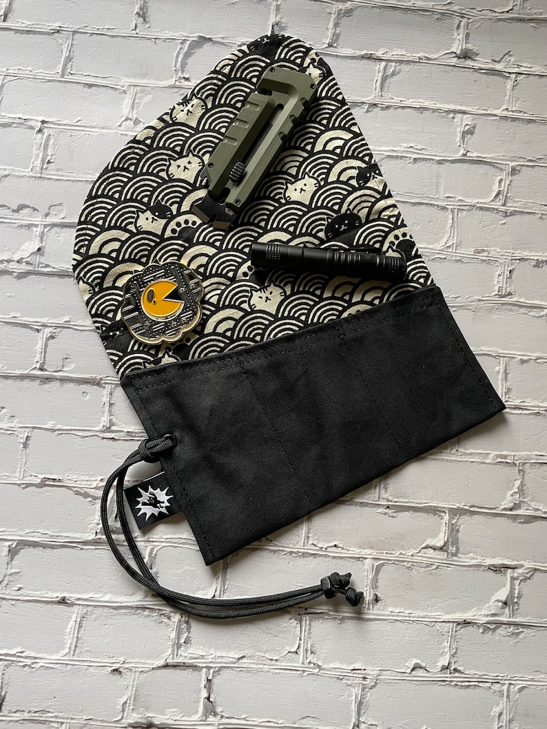 K&ouml;nnte beinhalten: Eine schwarze Stofftasche mit Kordelzug und einer kleinen Tasche auf der Vorderseite. Die Tasche hat drei F&auml;cher im Inneren. Die Tasche liegt auf einer wei&szlig;en Ziegelstein-Oberfl&auml;che. Die Tasche ist mit einem schwarz-wei&szlig;en Muster aus Wellen und Katzen verziert. Ein gr&uuml;nes Universalmesser und eine schwarze Taschenlampe liegen auf der Tasche.