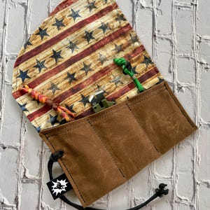 May include: A brown waxed canvas tool roll with a flap featuring a distressed American flag design. The roll is open, revealing a small green tool and two colorful lanyards. A black drawstring secures the roll.