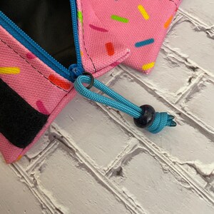 May include: A pink zippered pouch with a colorful sprinkle pattern. The pouch has a black zipper pull and a blue cord with a dark blue bead.