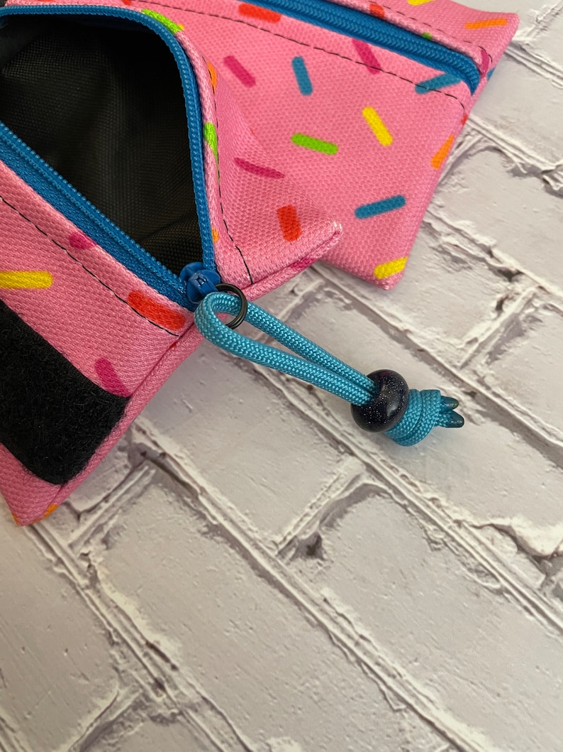 May include: A pink zippered pouch with a colorful sprinkle pattern. The pouch has a black zipper pull and a blue cord with a dark blue bead.