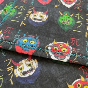 May include: Black fabric with a repeating pattern of colorful Japanese demon masks. The masks have different expressions and are surrounded by Japanese text. The text reads "元ホラー死" which translates to "Original Horror Death".