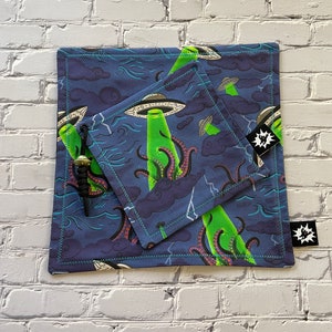 Op de afbeelding: Twee blauwe stoffen zakjes met een groen en paars alien- en octopusontwerp. Het kleinere zakje zit in het grotere zakje. Beide zakjes hebben een zwart sterlogo.