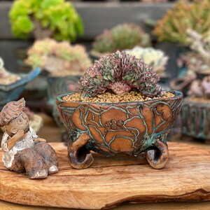 May include: A small succulent plant with pink and green leaves in a brown and blue ceramic pot with floral designs. The pot has three legs and is sitting on a wooden surface.