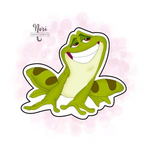 May include: A green frog cookie cutter with a wide smile and brown spots on its back. The cookie cutter is outlined in black and has the text "Nori Cookie Cutters by" at the top.
