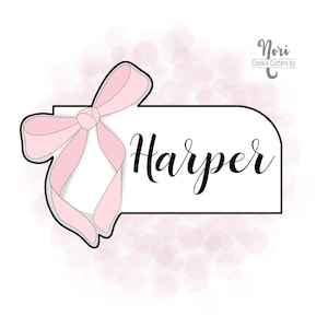 Puede incluir: Un cortador de galletas en forma de pancarta con un lazo rosa en el lado izquierdo. La pancarta dice "Harper".