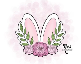 Puede incluir: Un cortador de galletas de orejas de conejo blanco con el interior rosa y hojas verdes alrededor de las orejas. Tres flores rosas se encuentran en la parte inferior del cortador. El texto "Nori Cookie Cutters by" está en la esquina inferior derecha de la imagen.