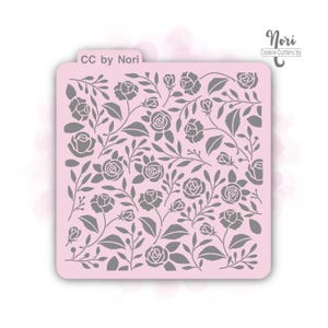 May include: A pink cookie stencil featuring a floral pattern of roses and leaves. The stencil has the text "CC by Nori" and "Nori Cookie Cutters by" in the upper corners. The design is ideal for decorating cookies and other baked goods.
