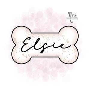 May include: A bone-shaped cookie cutter with the name "Elsie" in a cursive font. The cookie cutter has a cream-colored interior with gold flecks and a black outline. The background is a soft pink with blurred circles.
