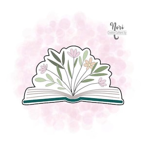 May include: A cookie cutter in the shape of an open book with a floral arrangement. The book is outlined in black with a teal spine and pages. The flowers are pink, peach, and white with green leaves. The text "Nori Cookie Cutters by" is in the upper right.