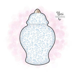 May include: A cookie cutter in the shape of a blue and white ginger jar with a lid. The jar has a floral pattern and a light pink background. The text "Nori Cookie Cutters by" is in the upper right corner.