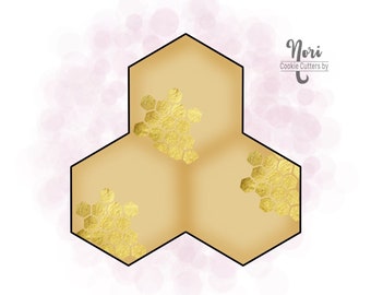 May include: A gold cookie cutter shaped like three connected hexagons with a honeycomb pattern. The cookie cutter is on a white background with pink watercolor circles.