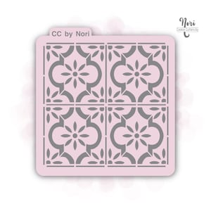 May include: A pink plastic cookie stencil with a repeating pattern of four gray squares with a floral design. The stencil is labeled "CC by Nori" in the top left corner and "Nori Cookie Cutters by" in the top right corner.