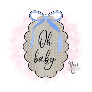 May include: A cookie cutter in the shape of a tag with a scalloped edge. The tag is gray with gold polka dots and the words "Oh baby" in black script. A blue bow and ribbons are at the top.